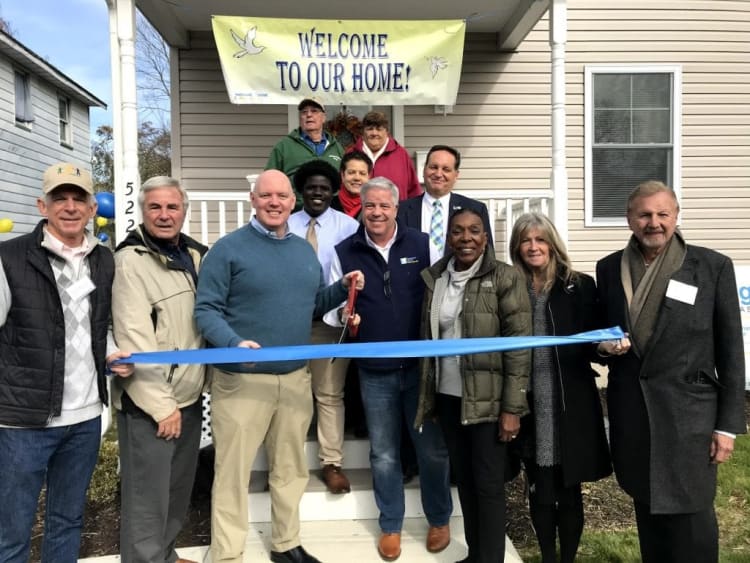 Group of people standing in front of a house holding a blue ribbon for a ribbon-cutting ceremony. A yellow banner above reads "WELCOME TO OUR HOME!" indicating a home dedication or community event. The setting is outdoors with a house porch and steps visible.