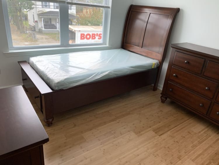 Dark wood bedroom set with a queen-sized sleigh bed featuring a plastic-covered mattress, matching dresser, and natural bamboo flooring.