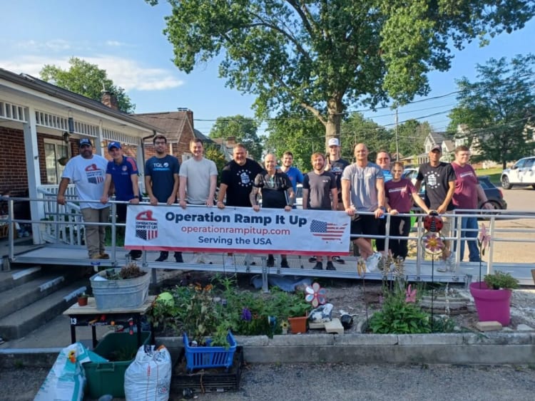 Group of people standing on a metal ramp holding a banner that reads "Operation Ramp It Up operationrampitup.com Serving the USA" in a residential neighborhood.