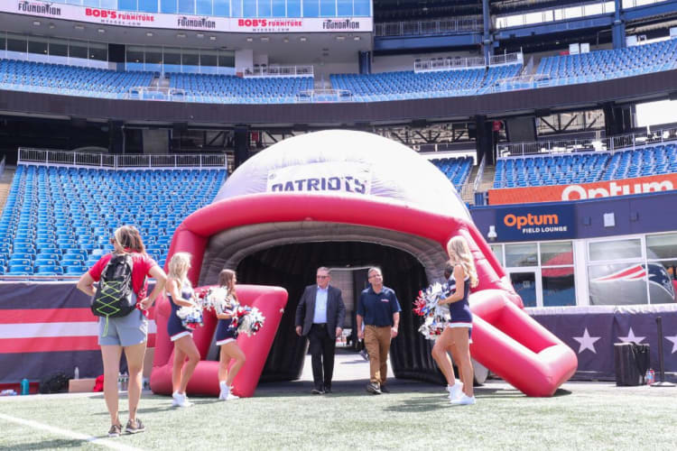 Two men walk out of a large inflatable Patriots tunnel on a football field, surrounded by cheerleaders holding pom-poms and stadium seating with blue chairs.