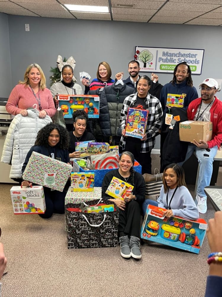 Group of people holding Fisher-Price and Play-Doh toys, gift boxes, and packages in a Manchester Public Schools room with a "Welcome Winter" sign.