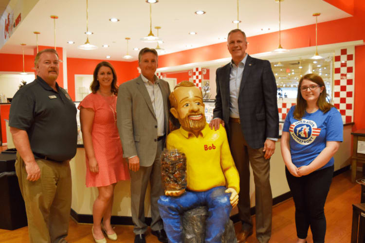 Five adults standing indoors around a seated statue wearing a yellow shirt labeled "Bob" holding a jar filled with candy, in a brightly lit room with red and white decor.