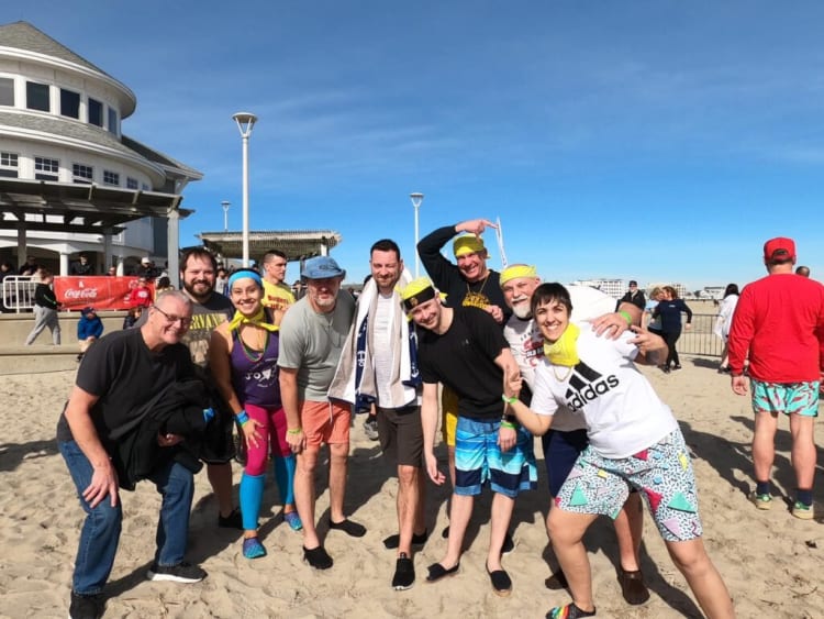 Group of people wearing colorful casual beachwear and bandanas posing on sandy beach near white building and clear blue sky.