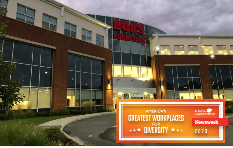 Bob's Discount Furniture store exterior with large windows and brick facade at dusk. Overlay text: "America's Greatest Workplaces for Diversity 2023" by Newsweek and Plant-A Insights Group.
