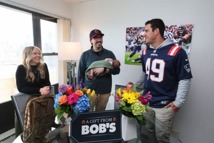 Three adults stand around a glass table with colorful floral arrangements and a sign reading "A GIFT FROM BOB'S." One man holds a baby wrapped in a green blanket. The man on the right wears a navy blue New England Patriots jersey with number 49. A football game poster is on the wall behind them.