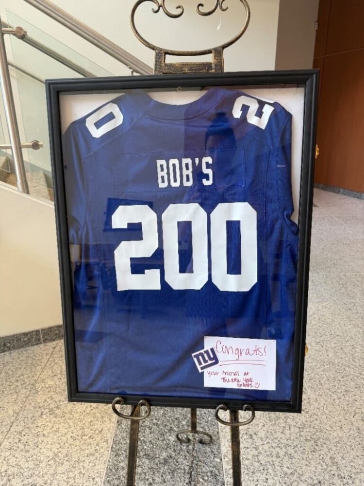 Framed blue New York Giants football jersey with number 200 and "BOB'S" on the back, displayed on a metal stand. Includes a note saying "Congrats! Your friends at the New York Giants."