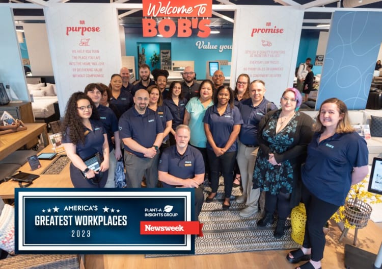 Team of employees in navy blue BOBS uniforms standing inside a modern furniture store with signs reading "Welcome to BOBS," "Our purpose," and "Our promise." Text includes: "America’s Greatest Workplaces 2023," "Plant-A Insights Group," and "Newsweek." The store features stylish, light wood furniture and home decor.