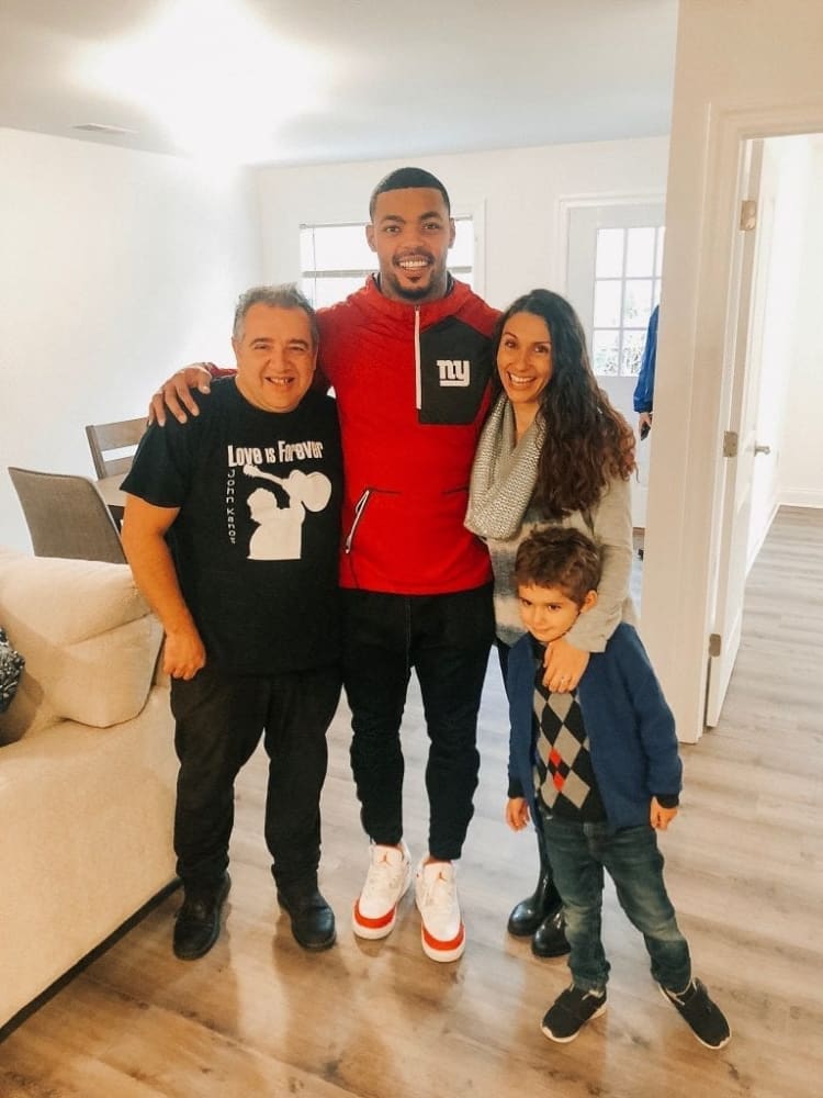 Four people standing together indoors on wood flooring, including a man in a red and black New York Giants jacket and white sneakers, a man in a black t-shirt that reads "Love is Forever," a woman wearing a gray scarf and black boots, and a child in a blue cardigan and argyle sweater.
