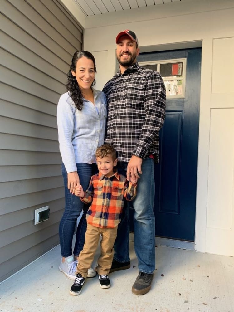 A family of three standing on a porch in casual fall attire: woman in light blue plaid shirt and jeans, man in black and white checkered shirt with jeans and cap, and child in orange and navy plaid shirt with tan pants and black sneakers.