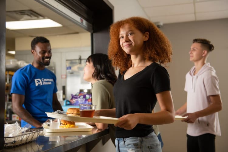Young people standing in line at a cafeteria counter holding trays with food, including sandwiches and drinks, in a casual indoor setting.