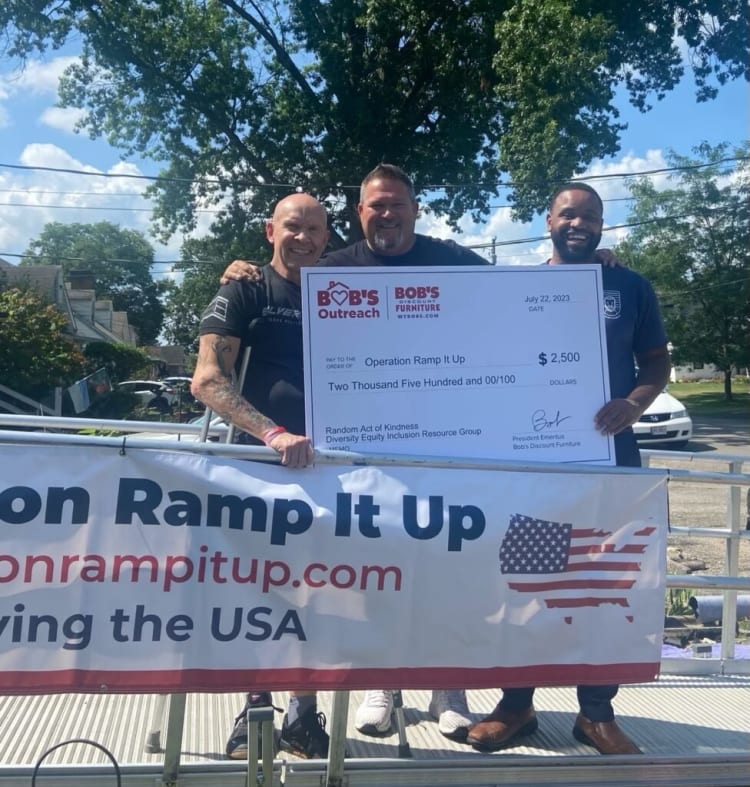 Three men holding a large $2,500 donation check from Bob’s Discount Furniture Outreach to Operation Ramp It Up, dated July 22, 2023, with a banner promoting onrampitup.com and supporting USA veterans.