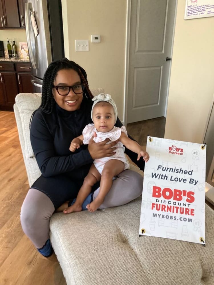 Person holding a baby sitting on a beige fabric sectional sofa next to a sign reading "BOB'S Discount Furniture Furnished With Love By MYBOBS.COM"