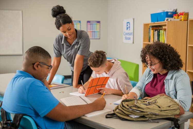 A group of students and a teacher collaborate at a classroom table with notebooks and pencils, promoting inclusive education.
