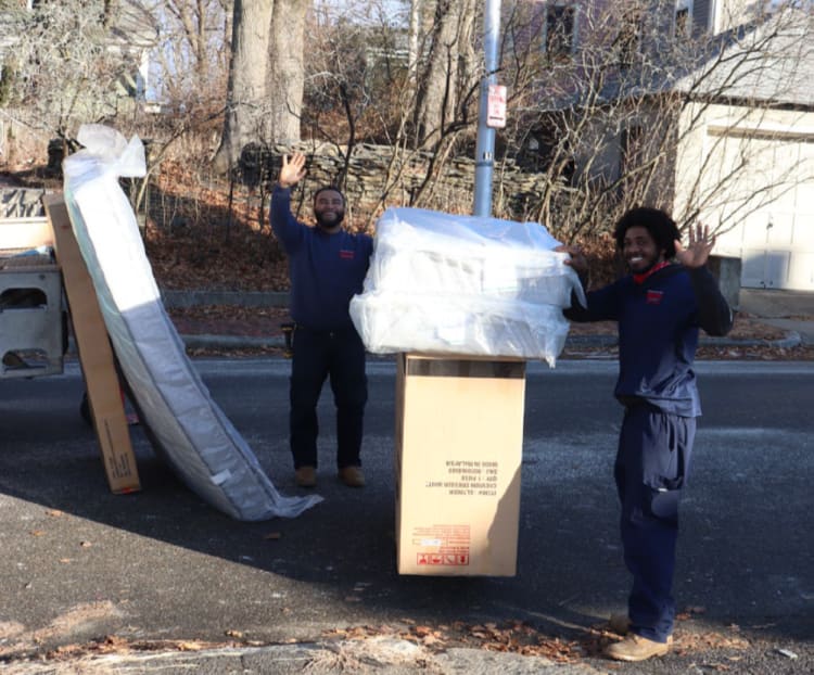 Two workers in navy blue uniforms stand on a street holding and waving near packaged mattresses and a large cardboard box labeled "Cushion Design Mattress, Made in Malaysia."