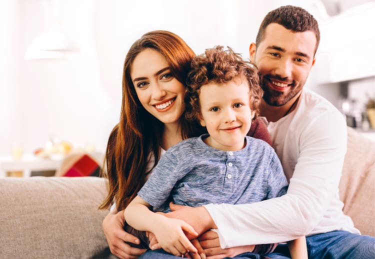 A family sitting closely on a beige couch in a bright living room, featuring a child in a blue henley shirt and adults in casual white and maroon tops.