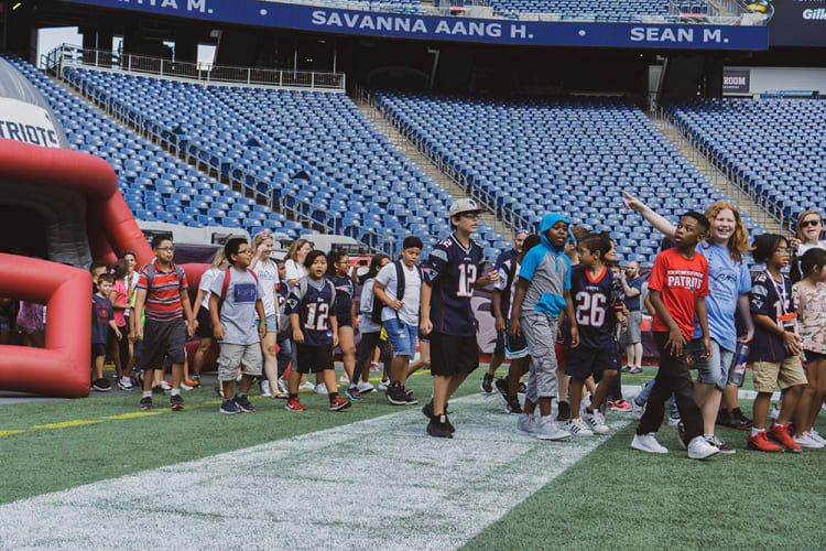 Children and adults walking on a football field near a large inflatable Patriots tunnel, wearing Patriots jerseys and casual clothing, with empty stadium seats in the background.