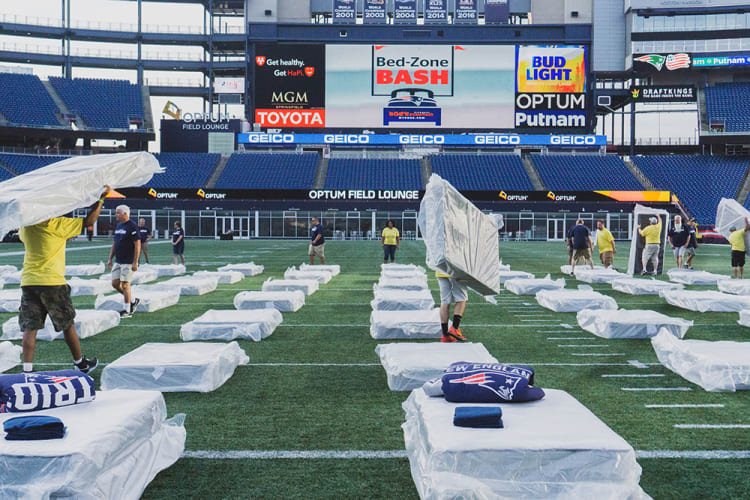 Workers set up covered beds with New England Patriots pillows on a football field at Optum Field Lounge, featuring large digital ads for Bed-Zone Bash, Bud Light, Toyota, and GEICO.