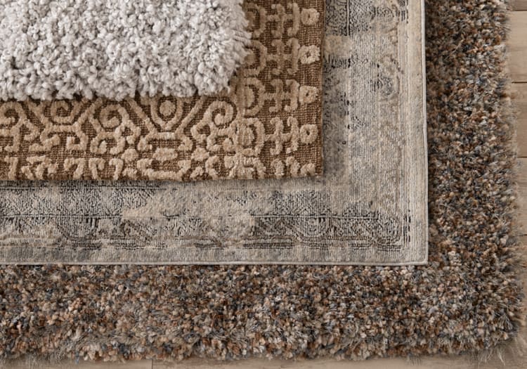 Stacked area rugs with varied textures and patterns: a beige shag rug on top, a brown rug with geometric designs in the middle, a vintage-style beige rug with faded floral borders, and a thick, multicolored shag rug at the bottom.