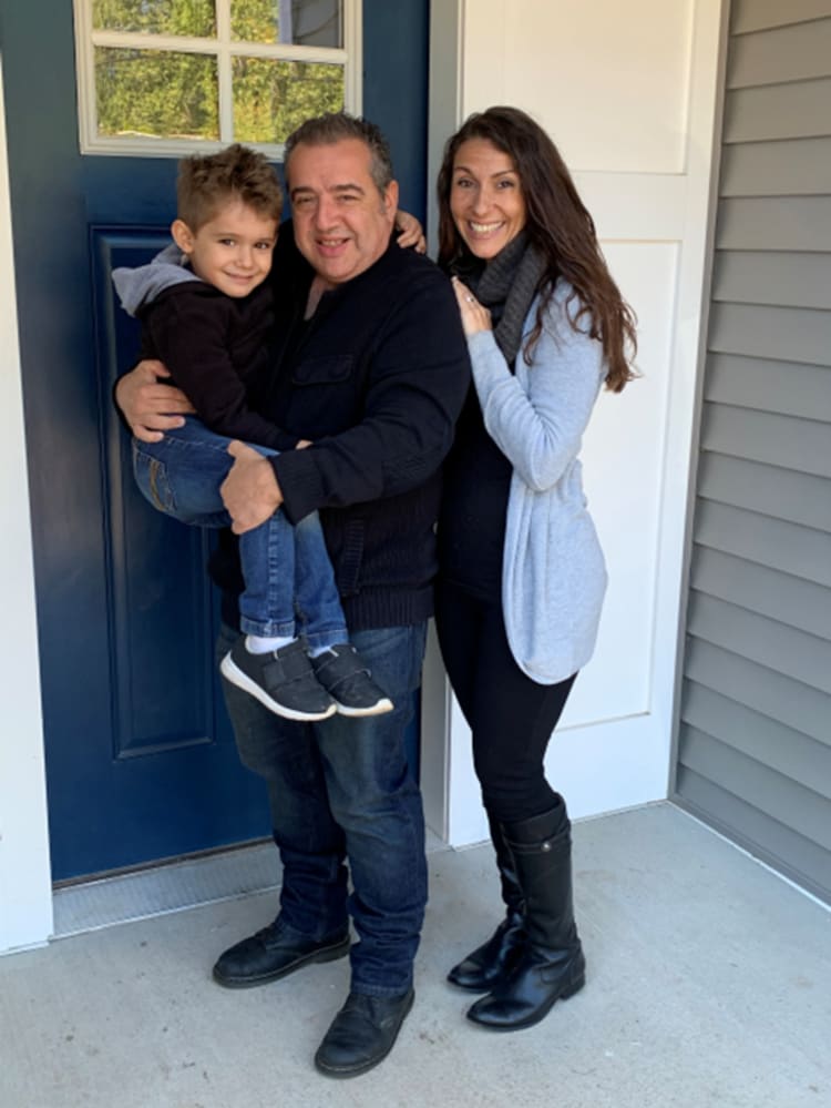 Family standing on a front porch with a blue door, dressed in casual fall clothing including jeans, black boots, and a gray cardigan.