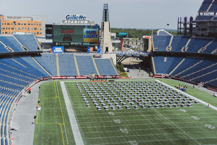 Gillette Stadium football field with rows of parked cars arranged in a grid on the turf, blue stadium seats, and digital scoreboard displaying "Bed Zone Bash" and "Bud Light" ads.