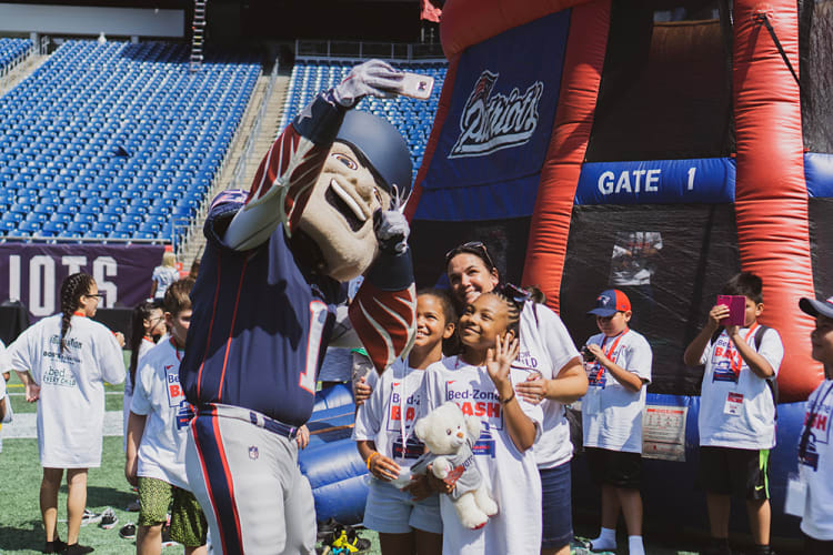 Mascot in New England Patriots uniform takes a selfie with children wearing white Bed-Zone Bash t-shirts at a stadium event near an inflatable Patriots-themed structure labeled "GATE 1."