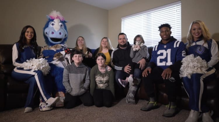 Group of people and a blue horse mascot sitting on a brown leather couch in a living room with a window, featuring two cheerleaders in blue and white uniforms holding white pom-poms.