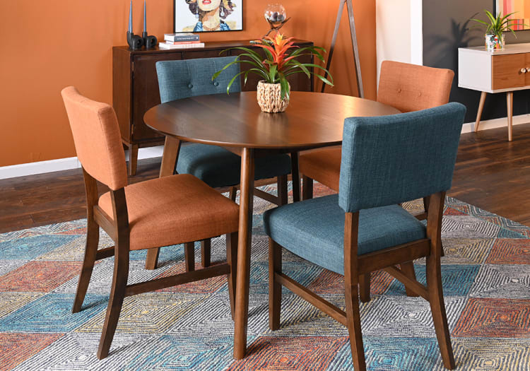 Round wooden dining table with a dark finish, surrounded by four upholstered chairs in burnt orange and teal fabric, set on a multicolored geometric rug in a modern dining room.
