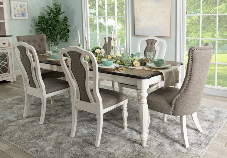 Dining room set with a rectangular wooden table featuring a dark top and white legs, surrounded by six upholstered chairs in gray fabric with white wooden frames, set on a patterned gray area rug near large windows.