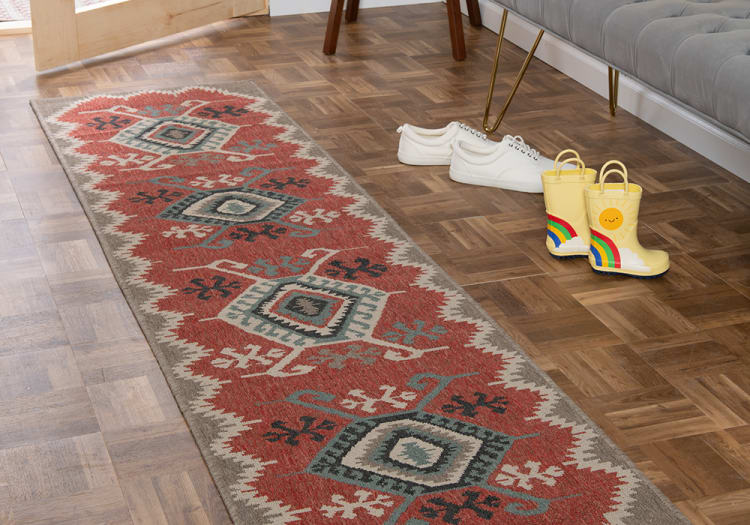 Long red and beige geometric patterned runner rug on wood parquet floor next to white sneakers and yellow rain boots with rainbow and sun design.