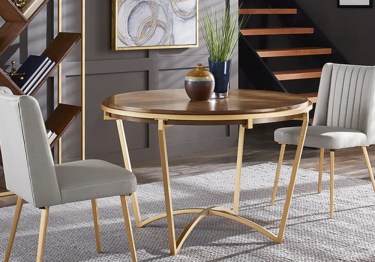 Round wooden dining table with gold metal frame and two gray upholstered chairs with gold legs on a textured gray rug in a modern room.