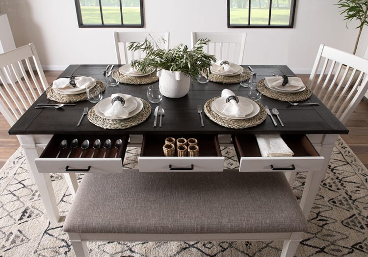 Dining table set with six white wooden chairs and a bench, featuring a dark wood top, woven placemats, white plates, cloth napkins, and open drawers with silverware and napkin rings.