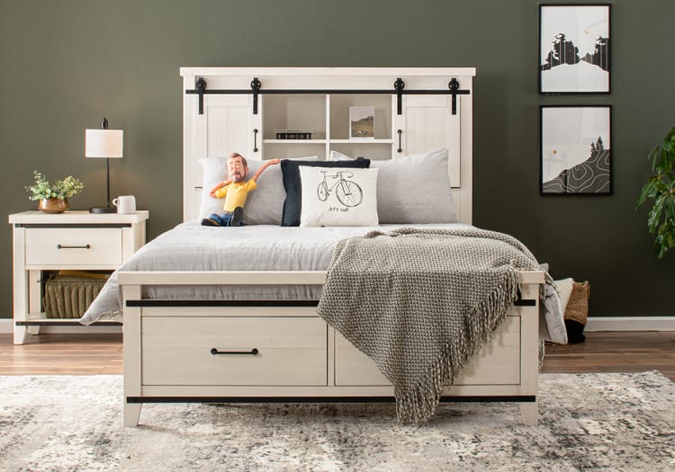 White wooden bed with storage drawers, gray bedding, textured throw blanket, decorative pillows including one with a bicycle and "let's ride" text, matching nightstand with lamp and plant, dark green wall with framed art and hardwood floor.