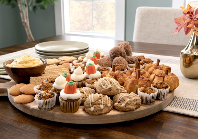 A wooden serving board with assorted fall-themed baked goods including pumpkin cupcakes with frosting, cinnamon rolls, pumpkin-shaped cookies, and various spiced treats, set on a dining table with stacked plates and a bowl of dip.