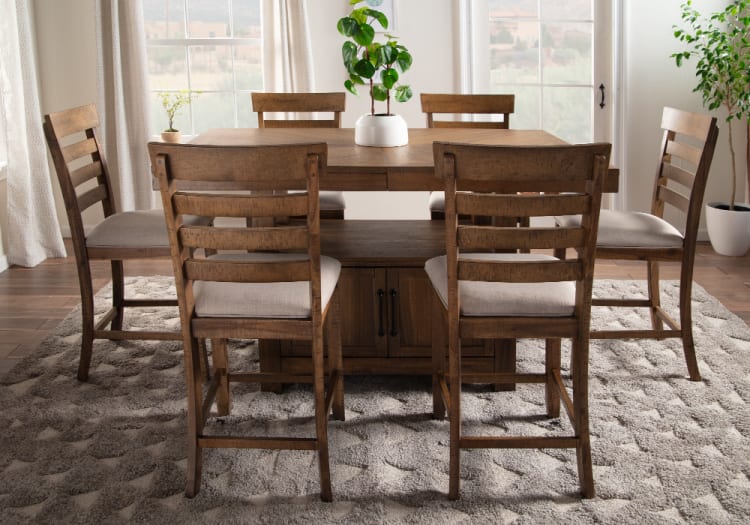 Rustic wooden dining table set with six ladder-back chairs featuring beige cushioned seats, placed on a textured gray rug in a bright room.