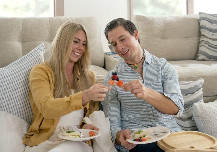 Two people sitting on a beige sofa, holding plates with sandwiches and fruit skewers, enjoying a casual indoor meal together.