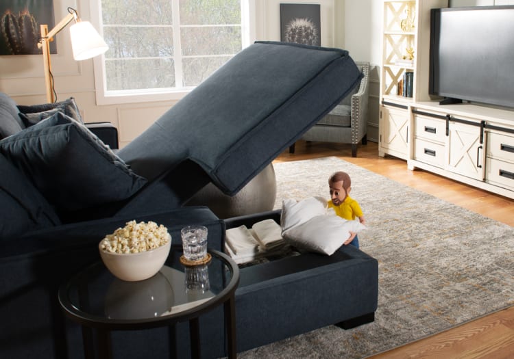 Dark blue fabric sectional sofa with a storage chaise open, revealing folded white linens and pillows inside, placed in a bright living room with a gray area rug and white entertainment center.