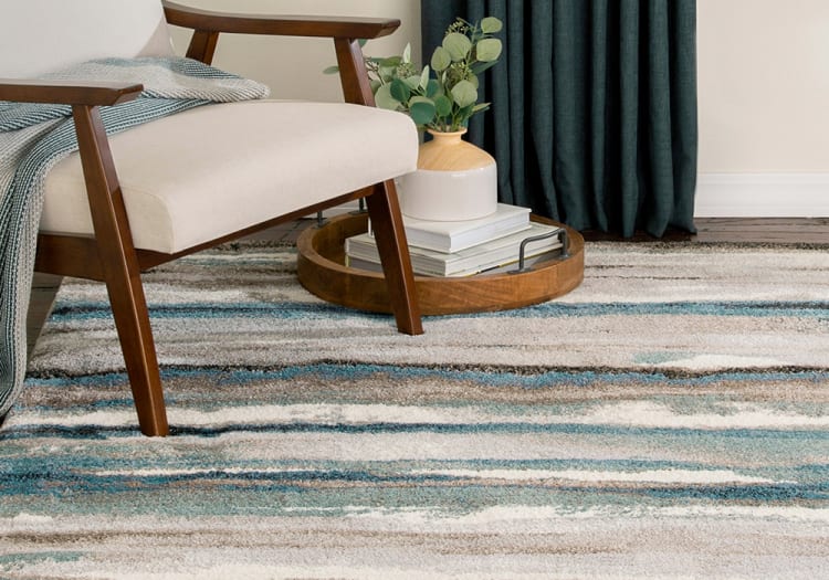 Mid-century modern beige upholstered armchair with wooden frame and angled legs on a striped blue, gray, and white textured area rug, next to a wooden tray holding a ceramic vase with green foliage and stacked books.