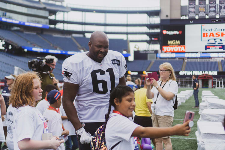 A football player in a white New England Patriots jersey with number 61 stands on a stadium field surrounded by fans, including children taking selfies and photos. Stadium signage displays brands like Toyota, MGM, and Gillette.