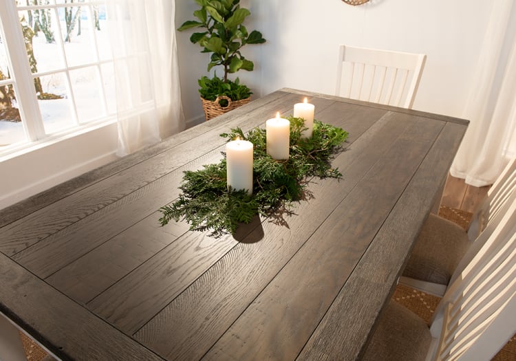 Rustic wooden dining table with three lit white pillar candles surrounded by green foliage centerpiece, set in a bright room with white chairs and natural light.