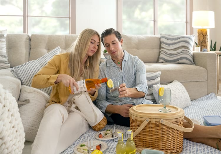 Two people sitting on a carpeted floor in a cozy living room, pouring a drink from a bottle with a lemon slice into a glass.