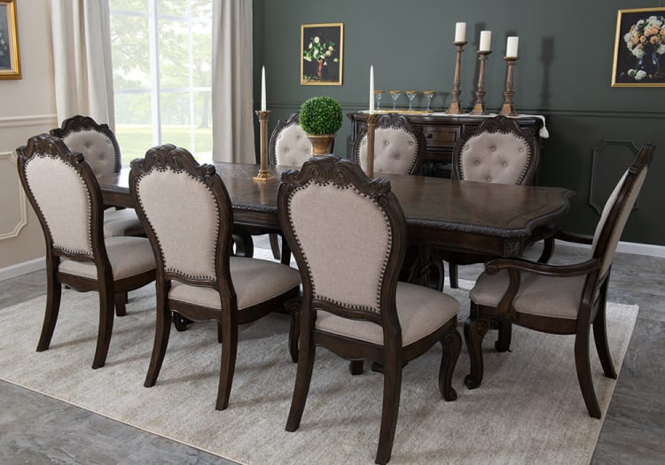 Dark wood dining table set with eight beige upholstered chairs featuring carved wood details, placed on a light area rug in a classic dining room.