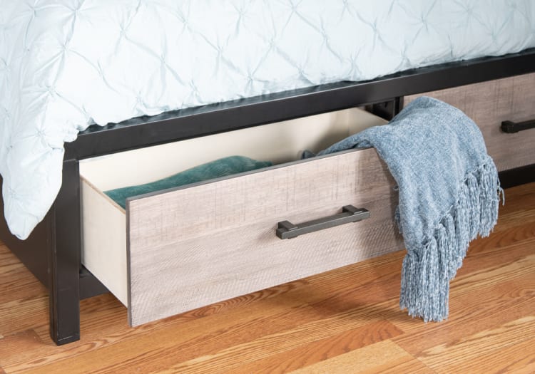 Under-bed storage drawer made of light wood with a black metal frame, partially open showing folded blankets and a blue throw with fringe on hardwood floor.