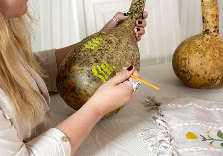 A person painting bright green leaf patterns on a large, brown, speckled gourd with a small paintbrush, showcasing handcrafted decorative art.