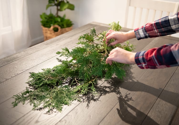 Hands arranging fresh green cedar branches on a wooden table, illustrating holiday wreath or centerpiece decoration preparation.