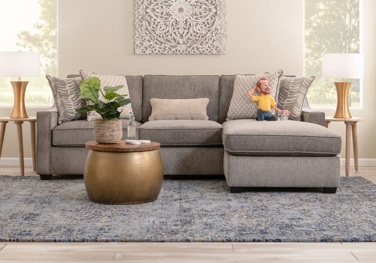Gray sectional sofa with patterned throw pillows, beige lumbar pillow, brass round coffee table with wooden top, green potted plant, and matching brass table lamps.