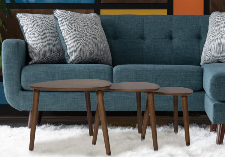 Teal fabric sectional sofa with tufted backrest and patterned white-gray cushions, paired with three nested wooden coffee tables on a white shag rug.