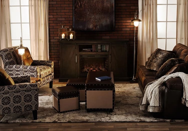 Cozy living room with brown leather sofa, patterned armchairs, beige and brown ottomans, brick wall, and warm lighting for a rustic ambiance.