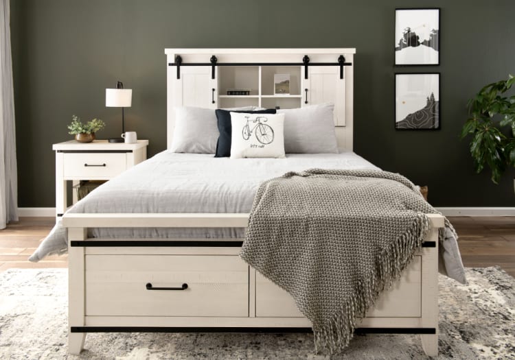 White wooden bed frame with built-in storage drawers, gray bedding, decorative pillows, and a textured throw blanket in a modern bedroom setting.