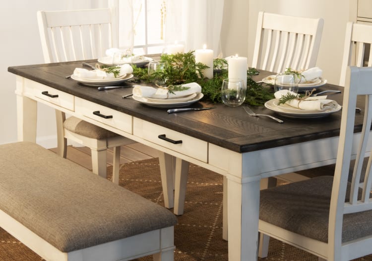 Dining table set with a dark wood top and cream-colored base, featuring white plates, rolled napkins with greenery, candles, and a cushioned bench and chairs.