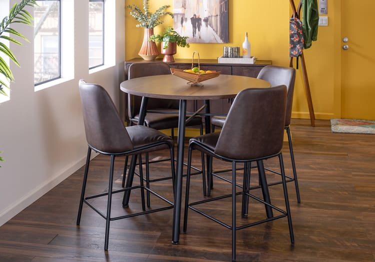 Modern dining area with a round wooden table and four brown leather cushioned chairs with black metal legs on dark hardwood flooring, set against a yellow wall with decorative plants and artwork.